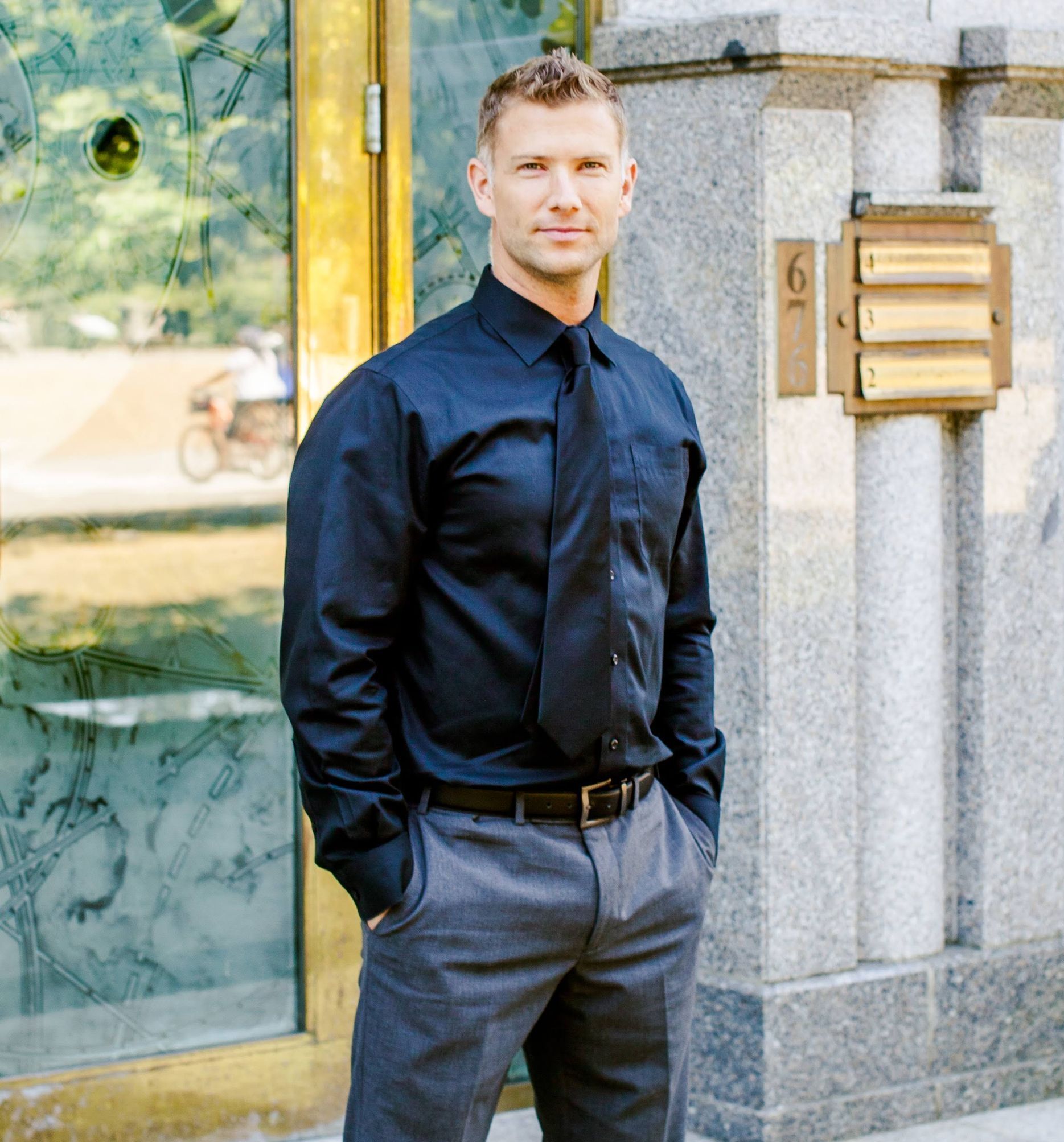 A man in a black shirt and tie is standing in front of a building with the number 6 on it