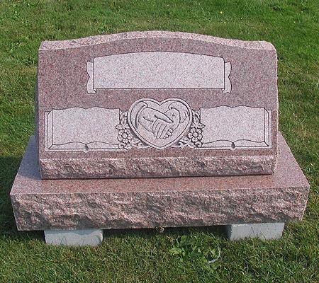 Red granite headstone with heart emblem, two clasped hands, rectangular inscription areas, on a grassy plot.