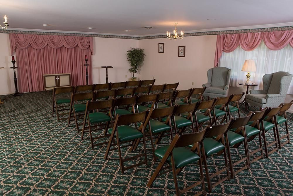 Funeral home interior with rows of chairs facing an empty space, pink curtains, green carpet.