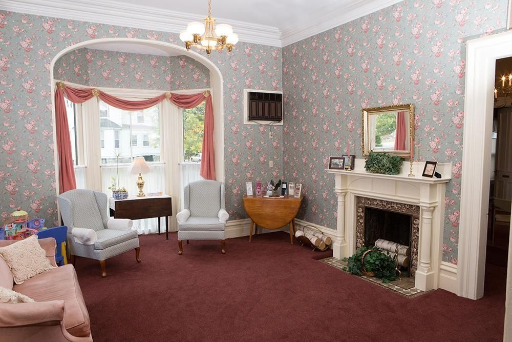 Room with floral wallpaper, a fireplace, two armchairs, a sofa, and a window with red drapes.