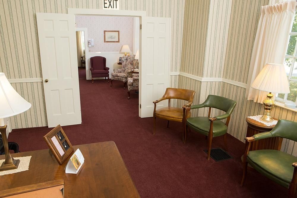 Waiting room with two chairs, desk, and view into another room with furniture. Exit sign above doorway.