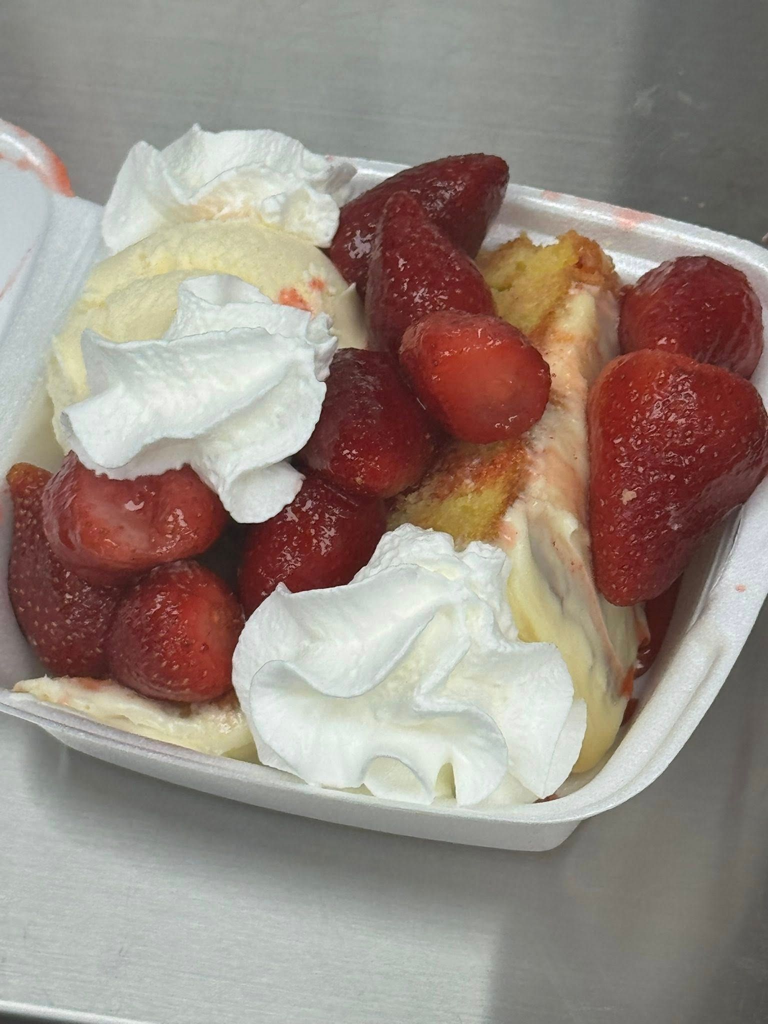 Strawberry shortcake dessert in a white takeout container, topped with whipped cream and fresh strawberries.
