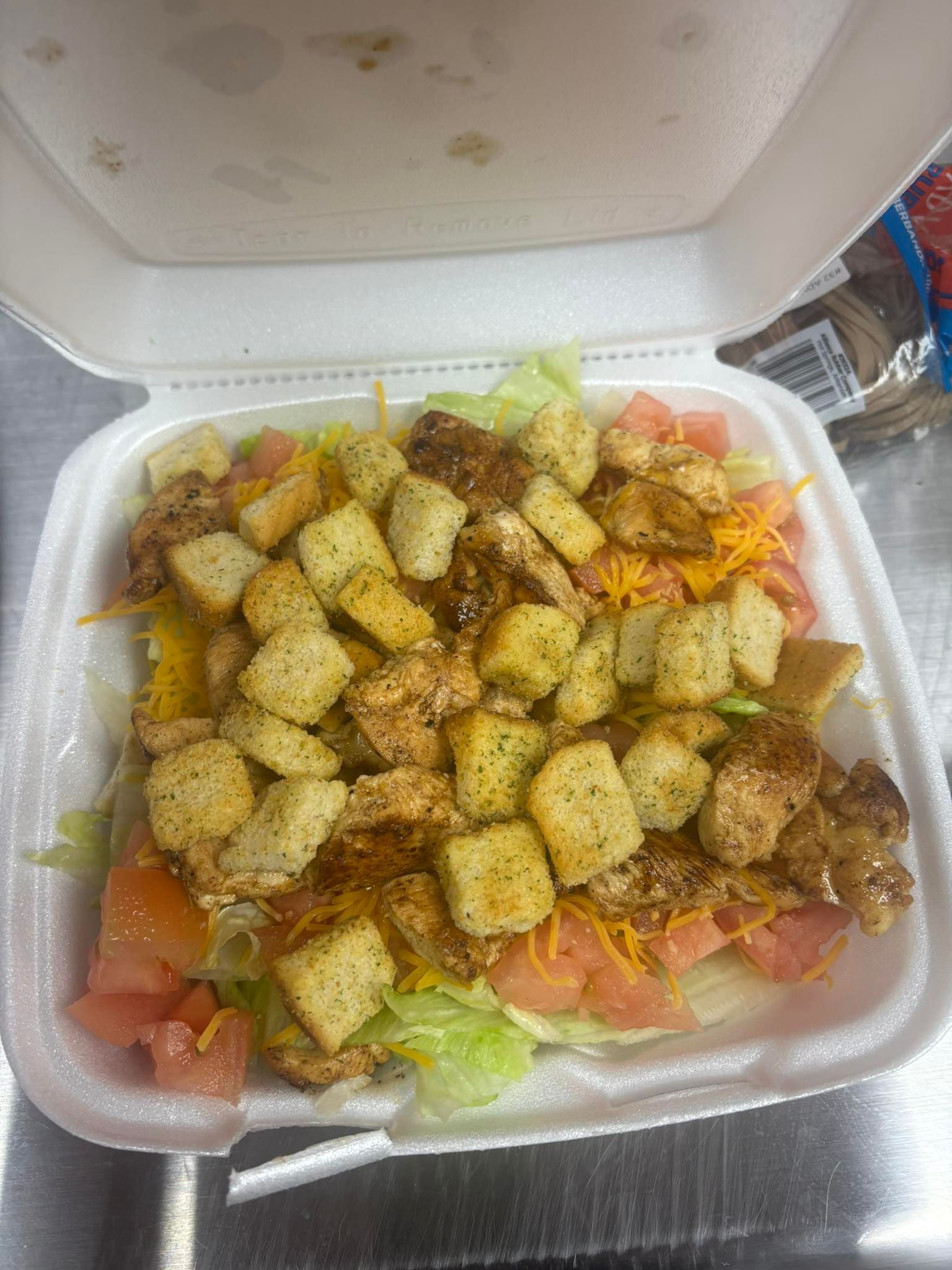 Chicken salad in a white takeout container.  It includes lettuce, tomato, cheese, croutons and cooked chicken.