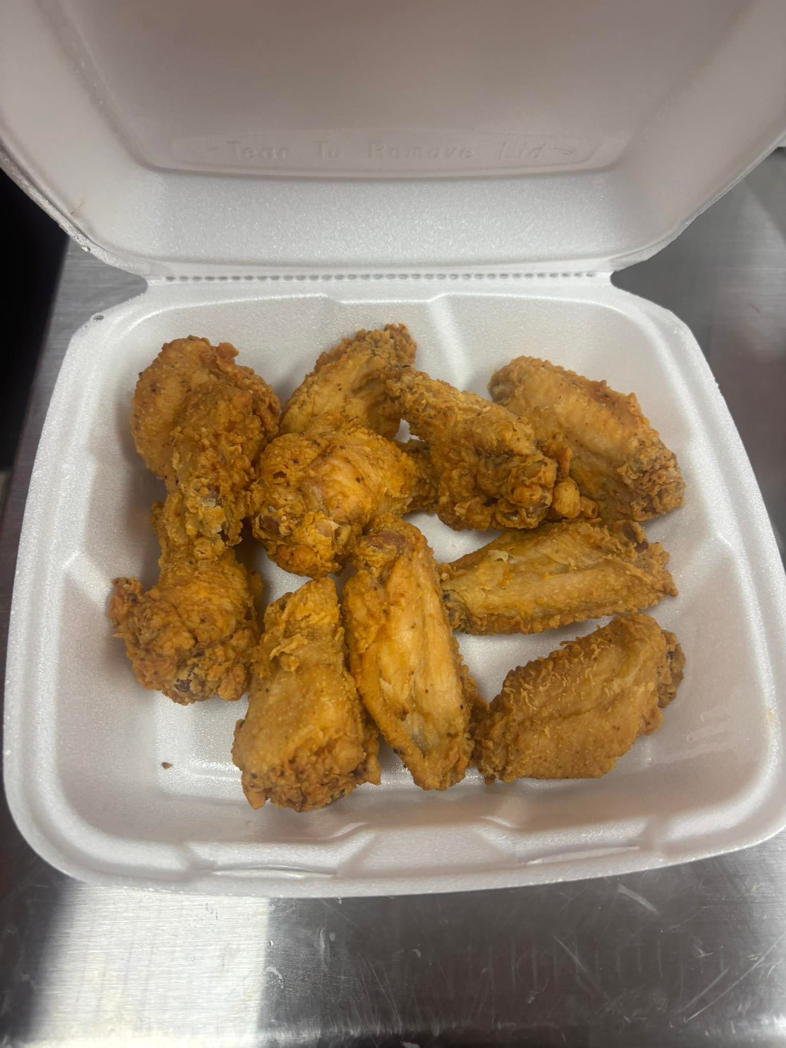 A container of fried chicken wings, golden brown and seasoned, arranged in a styrofoam container on a silver surface.