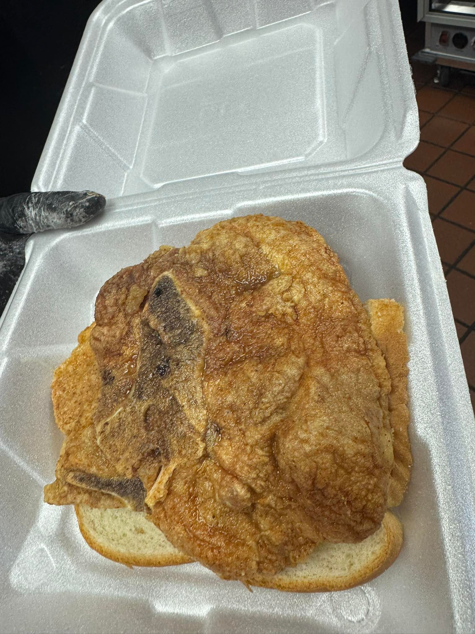 A fried pork chop sandwich sits inside a white styrofoam container. The chop is golden brown, resting on a slice of bread.