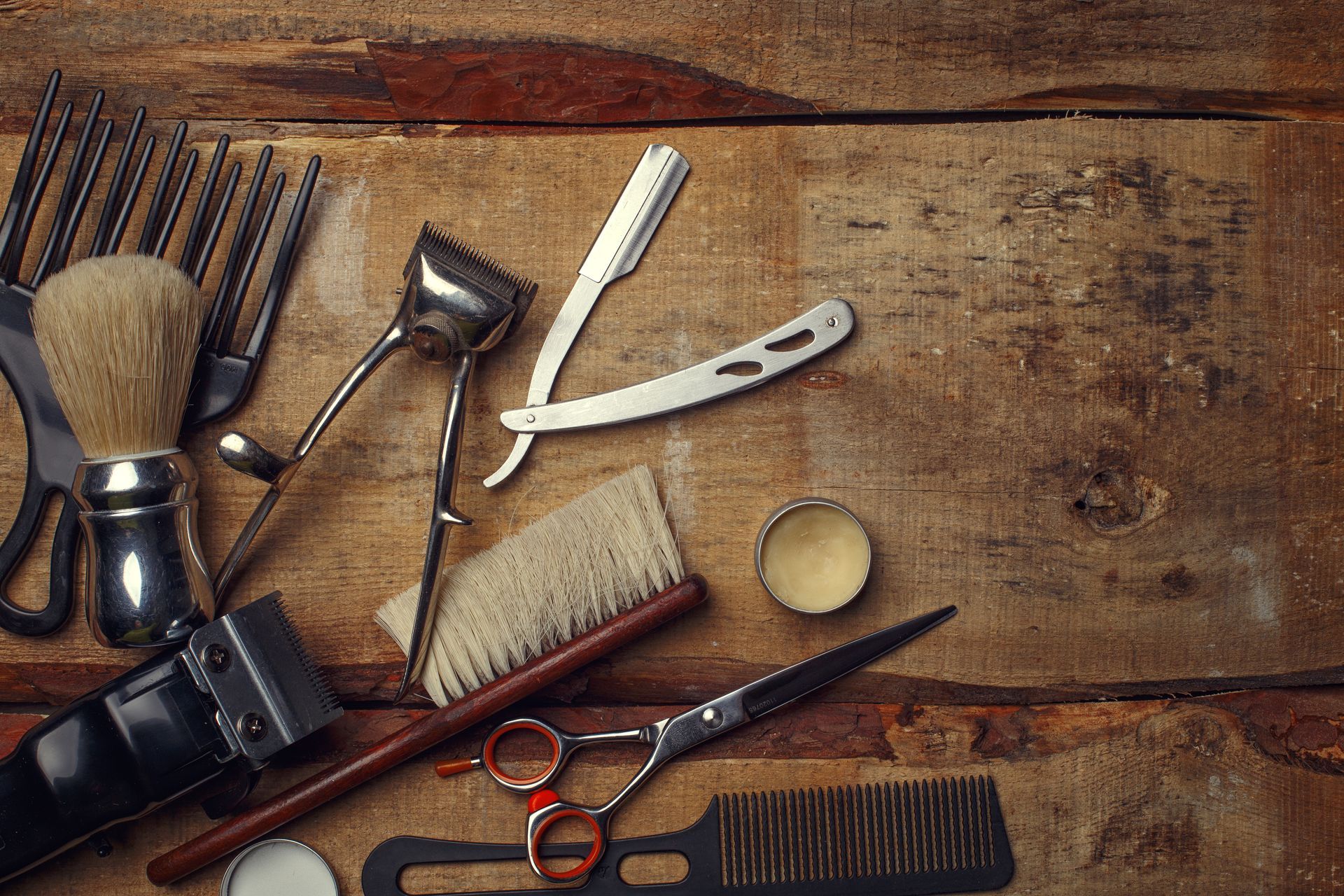 Barber tools, including clippers, razor, scissors, and brushes, on a wooden surface.