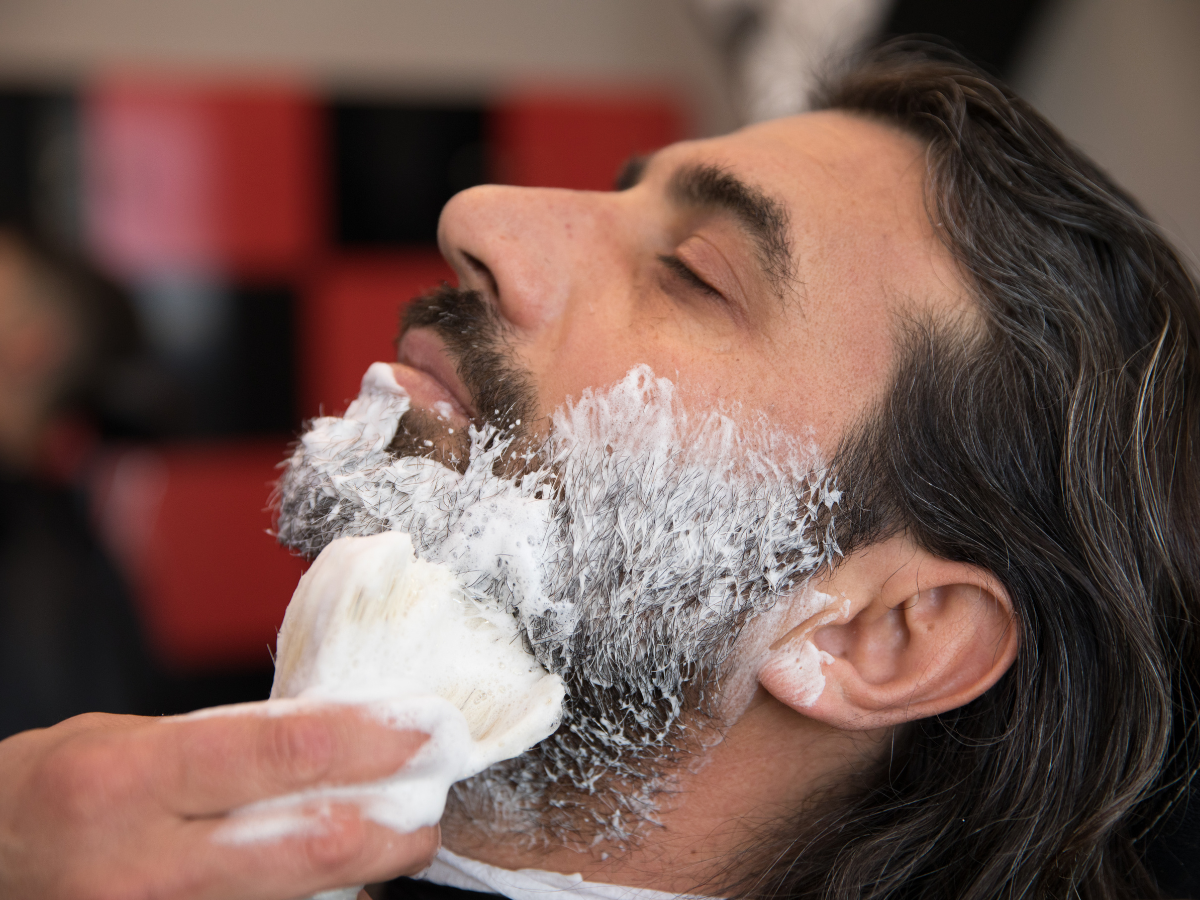 Man getting a shave at a barber shop. A barber applies shaving cream to his beard with a brush. Man getting a shave at a barber shop. A barber applies shaving cream to his beard with a brush.