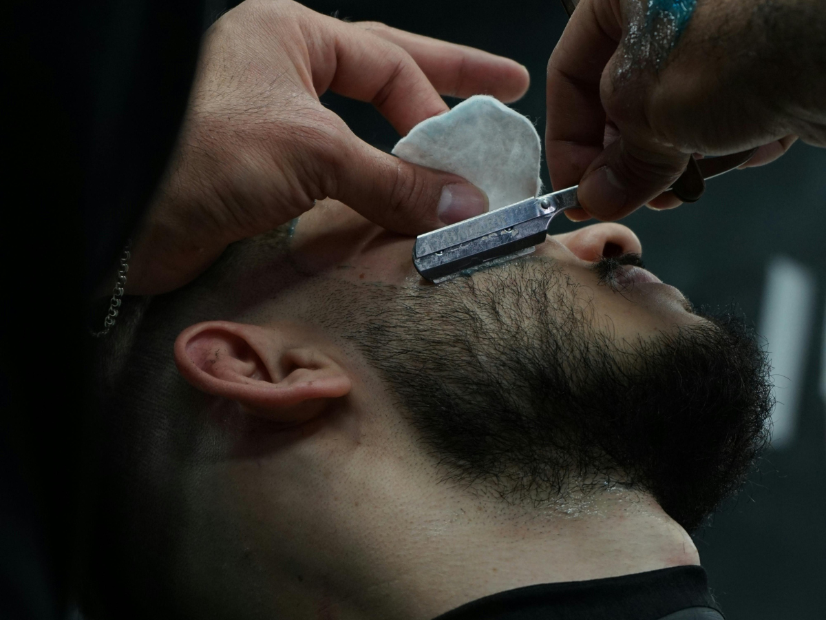 Barber shaving a man's face with a straight razor, using a cotton pad.