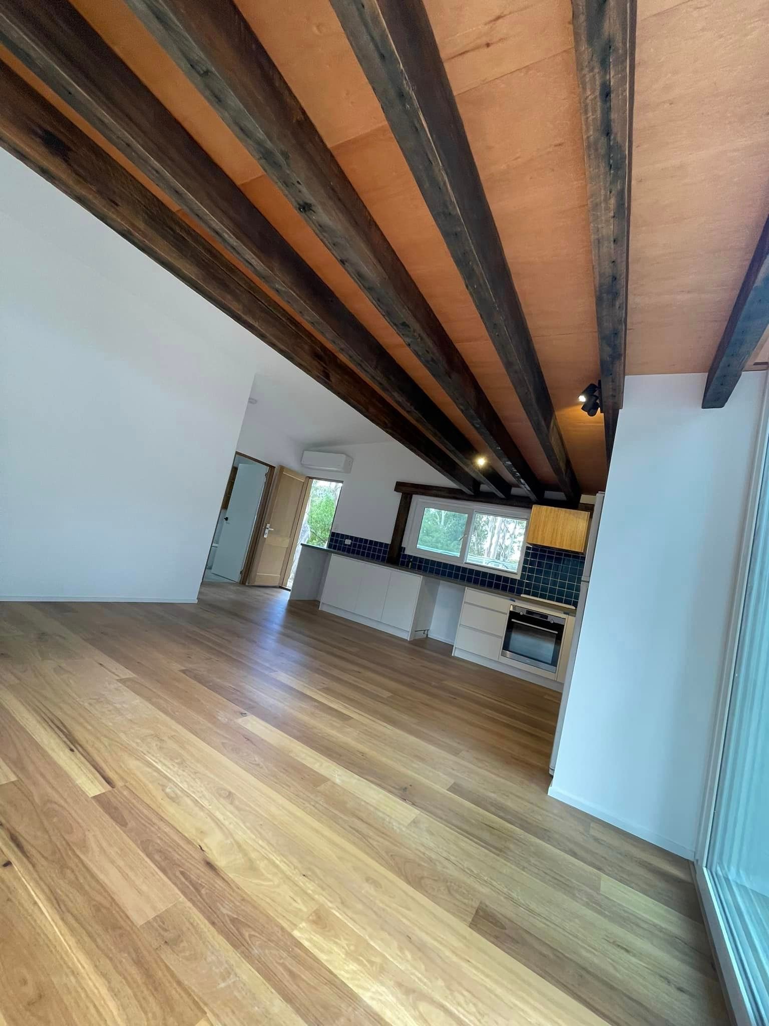 Wooden-floored living area with exposed beams, leading to a kitchen with white cabinets, and an open door.