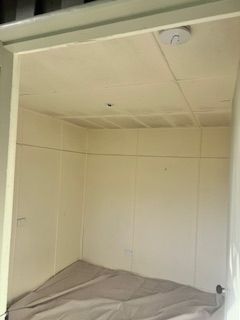 Small, empty room with beige walls and a bed covered by a sheet. A smoke detector is on the ceiling.