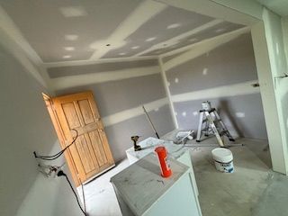 Room under construction, drywall on walls and ceiling. Wooden door, white cabinet, tools, and a ladder are visible.
