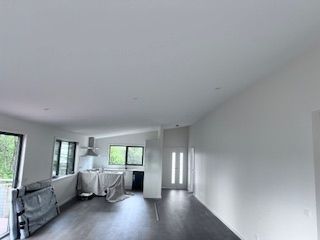 Empty interior of a room with white walls, dark floor, and windows. Kitchen area visible.