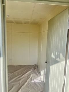 Interior view of a small, empty, cream-colored shed with a partly open door and a sheet on the floor.
