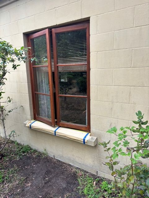 Wooden-framed window open against a beige, textured wall. A wooden beam is attached below, with a garden around.