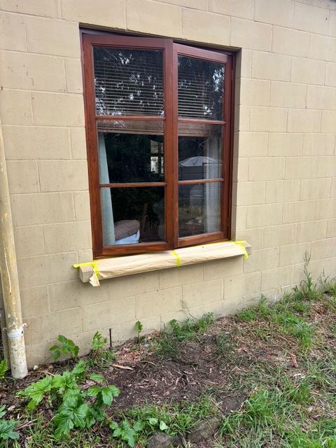 Brown-framed window on a tan brick wall. Yellow tape covers the sill. Green plants grow below.