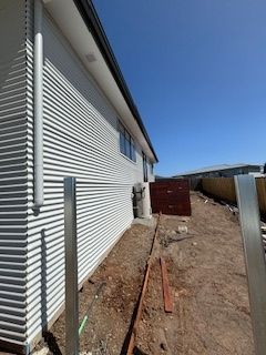 Side of a house with corrugated metal siding, metal fence posts, and a dirt path under a clear blue sky.