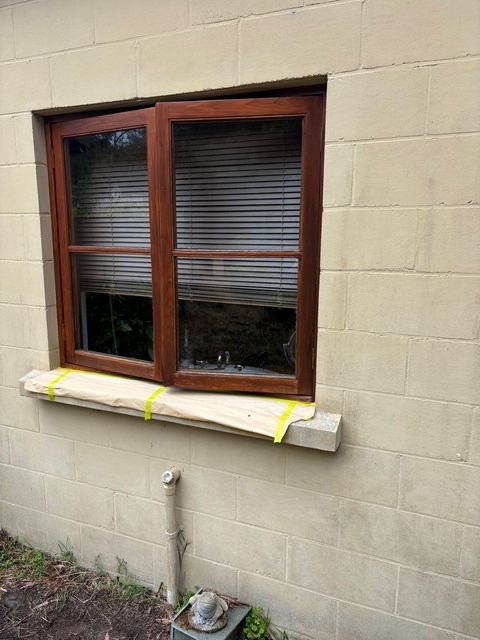 Window in a tan brick wall; wood frame, closed blinds, wood plank taped to window ledge.