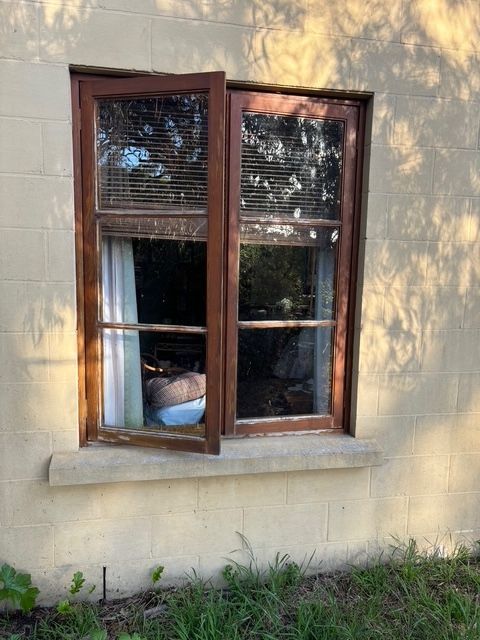 Brown-framed window, one side open, on a light yellow building. Blinds and a curtain visible inside.