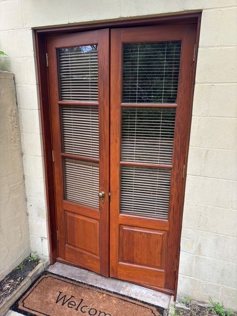 Double wooden doors with blinds and a welcome mat on a concrete wall.