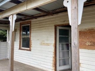 Exterior of a weathered, white-painted wooden house with a porch. A window and door are visible.