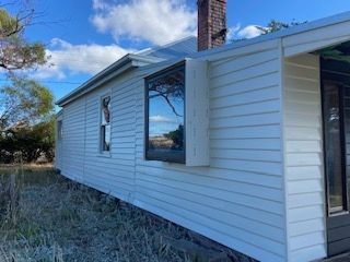 Side view of a white clapboard house with a large window reflecting the sky, set in a grassy area.