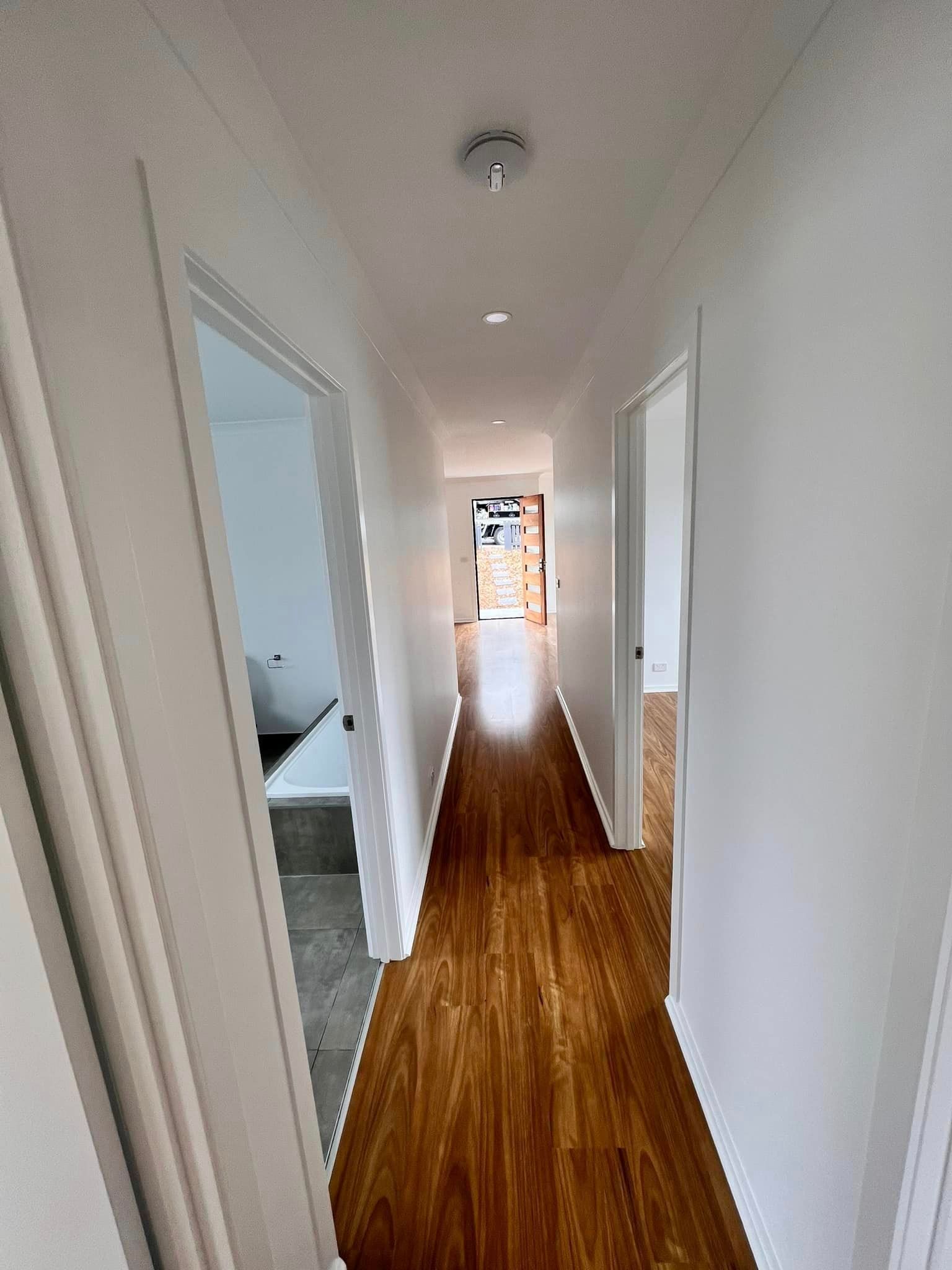 Narrow hallway with wooden floor, white walls and doors, leading to an open doorway.