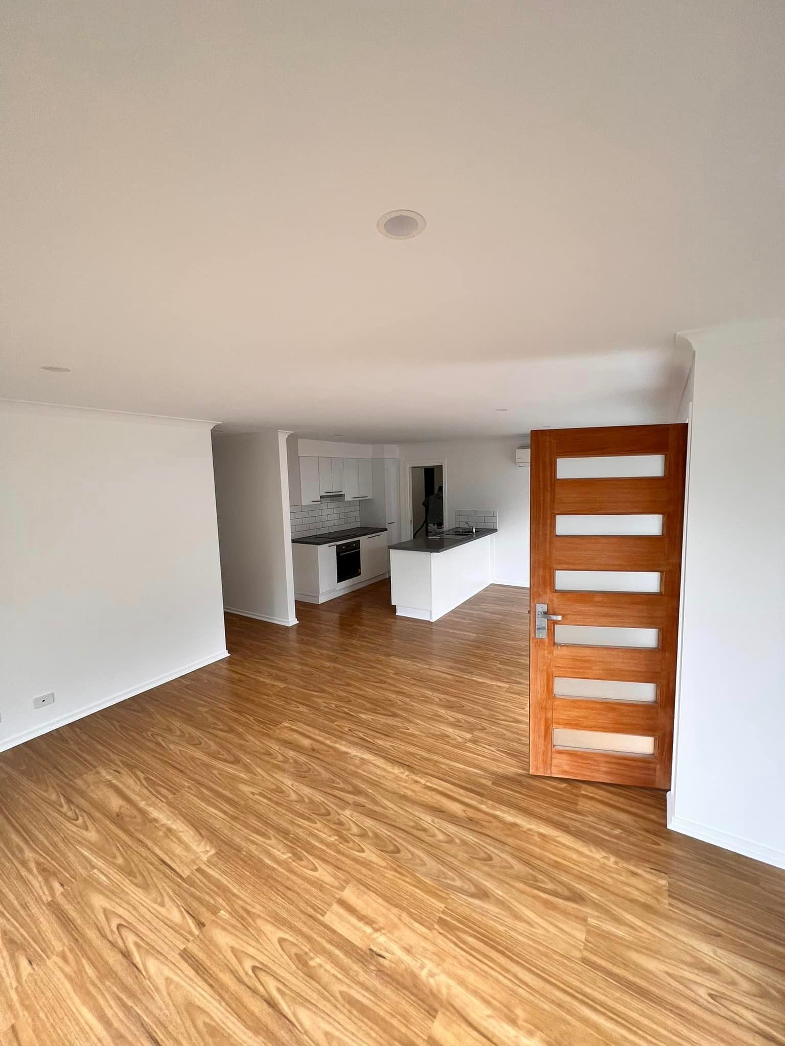 Open-plan living space with wood floors, white walls, and a view into the kitchen area. A wooden door is open on the right.