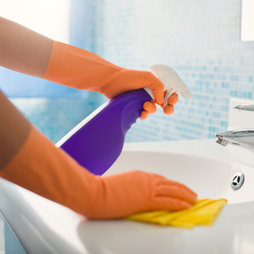 A person wearing orange gloves is cleaning a sink with a spray bottle