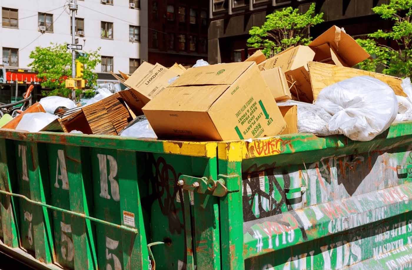 A green dumpster filled with cardboard boxes and plastic bags
