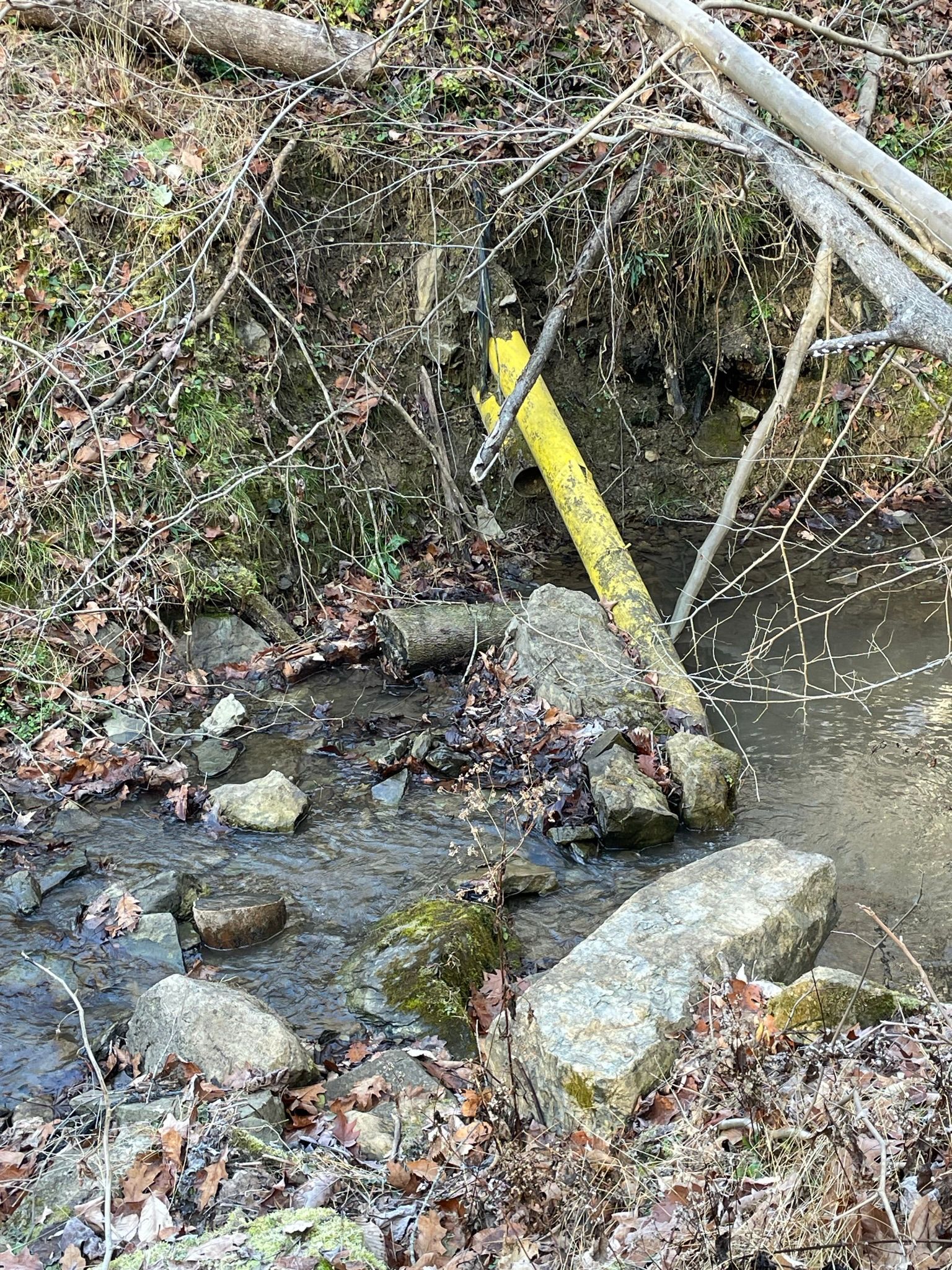 Close-up of a damaged yellow steel natural gas pipeline hanging over an eroded streambank, partially submerged in a rocky creek.
