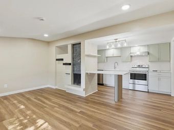 Bright empty kitchen and living area with hardwood floors, white cabinets, and a breakfast bar