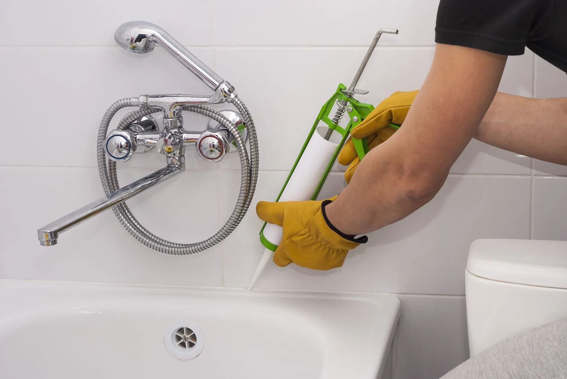 Person sealing the edge of a bathtub with a caulk gun. They're wearing work gloves.