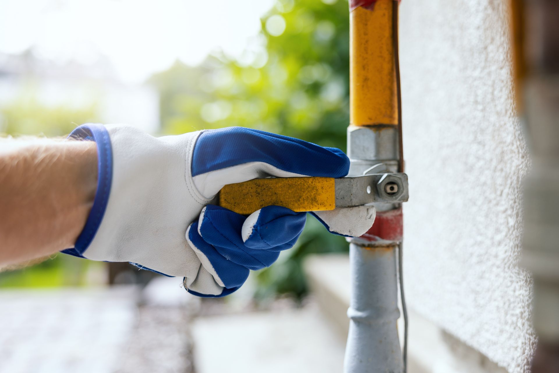 Hand in a blue and white glove turning a yellow gas shut-off valve on a pipe outdoors.