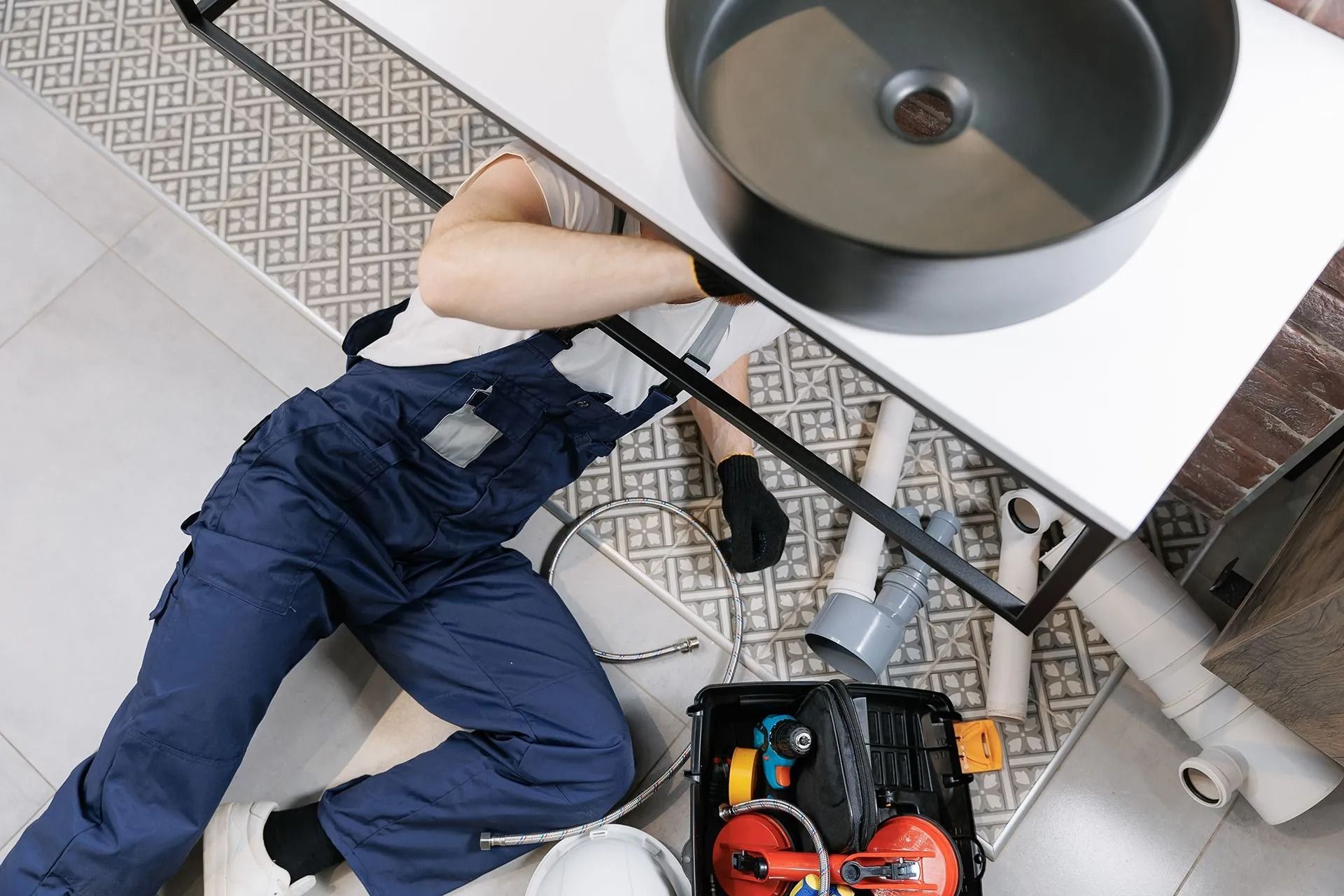 Plumber in blue overalls under a bathroom sink, working on pipes, tools and toolbox visible.