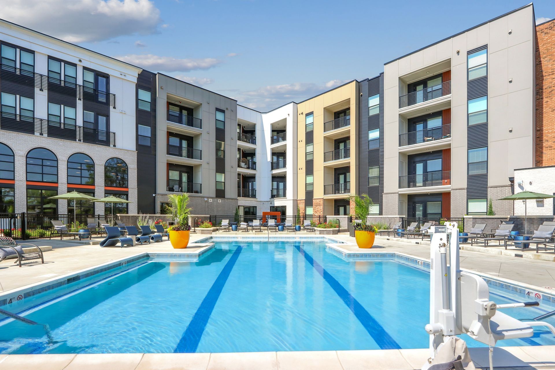 People working at a large wooden table in a bright office; two using laptops, one writing, plants and supplies on the table.