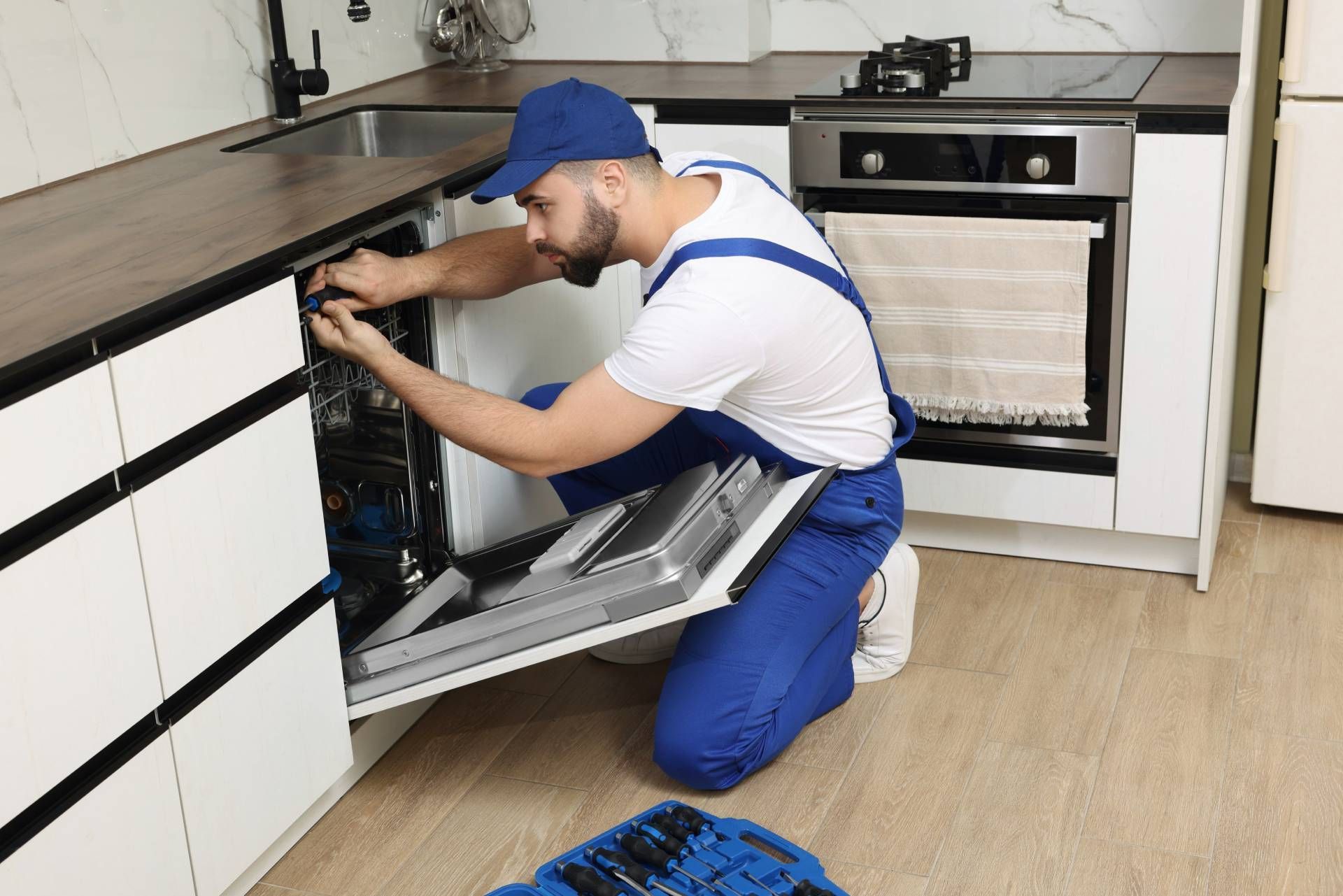 A repairman kneels to fix a dishwasher in a modern kitchen.