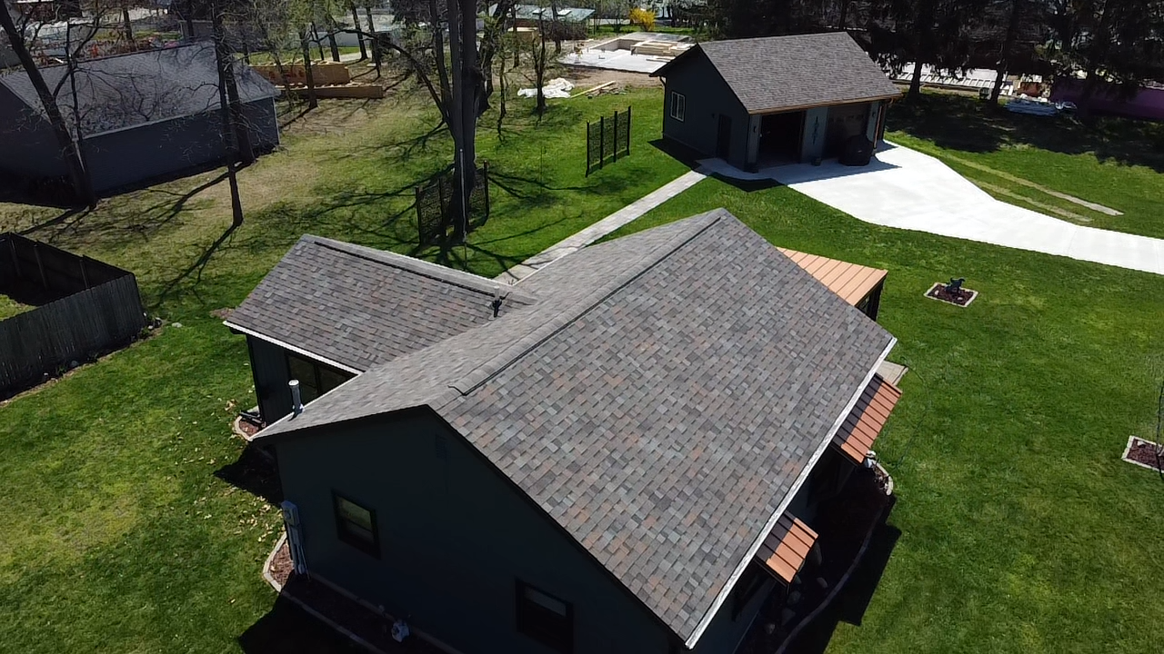 Aerial view of a gray house with a brown roof, surrounded by green grass and a small outbuilding.