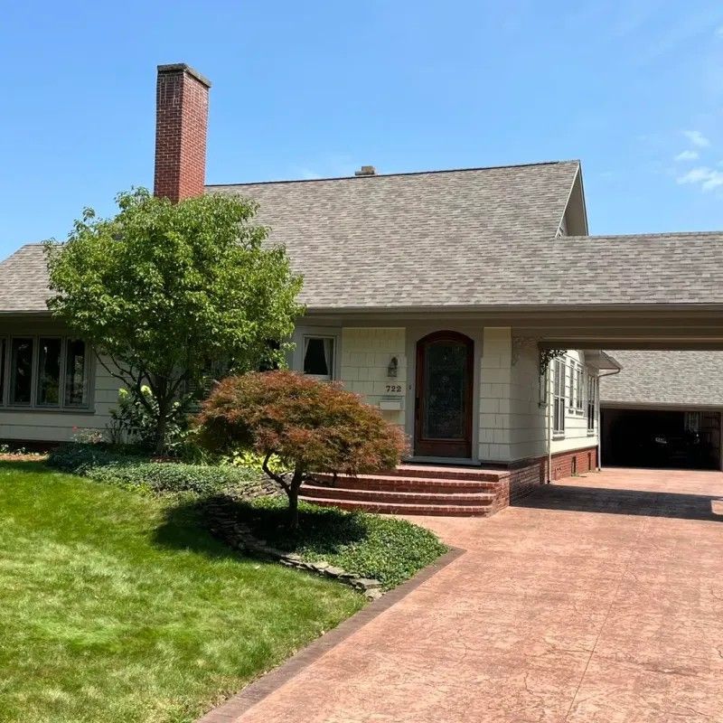Curb appeal of a cream-colored house with a red brick chimney, carport, and walkway. Lush green lawn and blue sky.