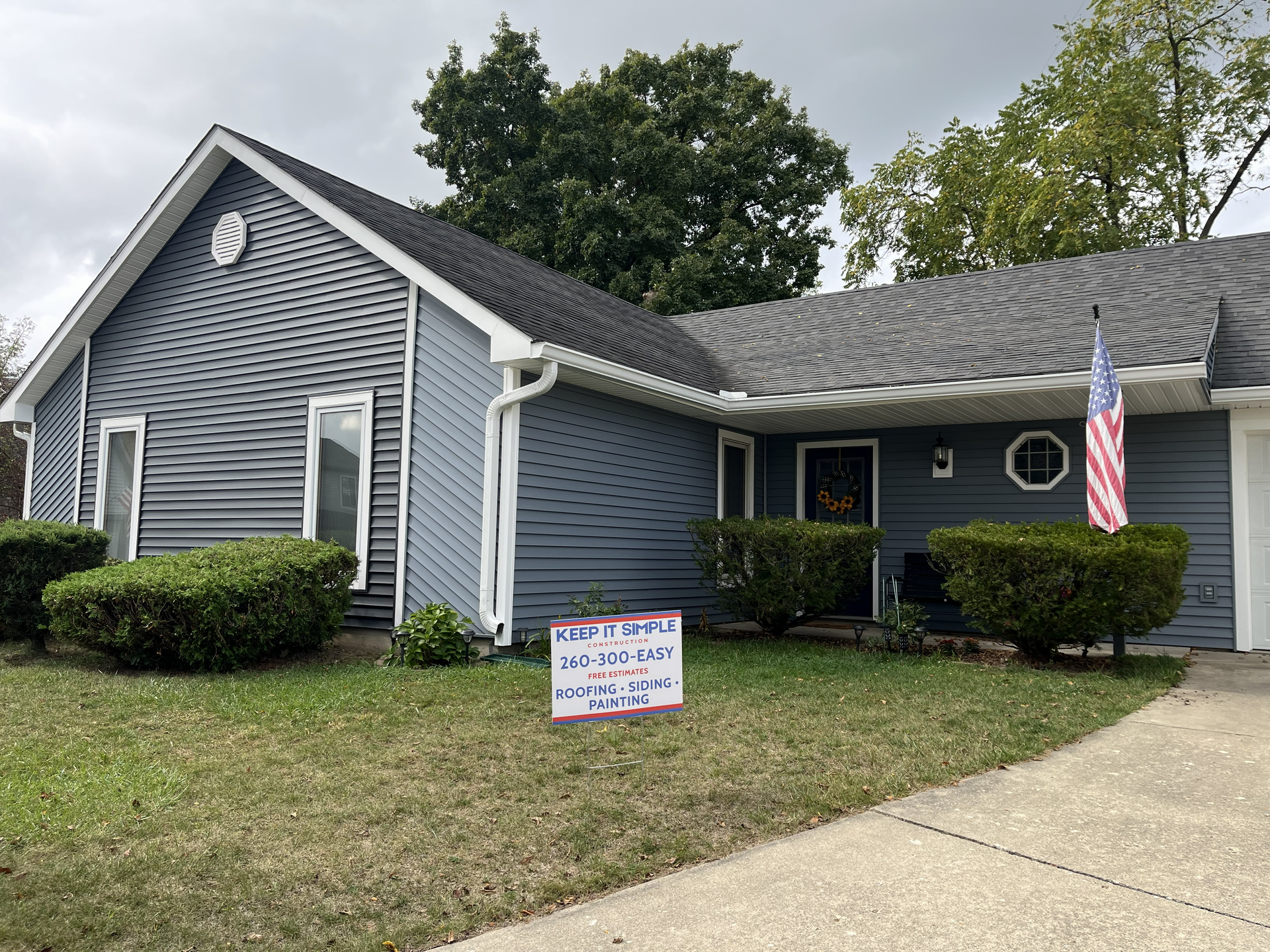 Blue house with bushes, American flag, and a sign on the lawn.