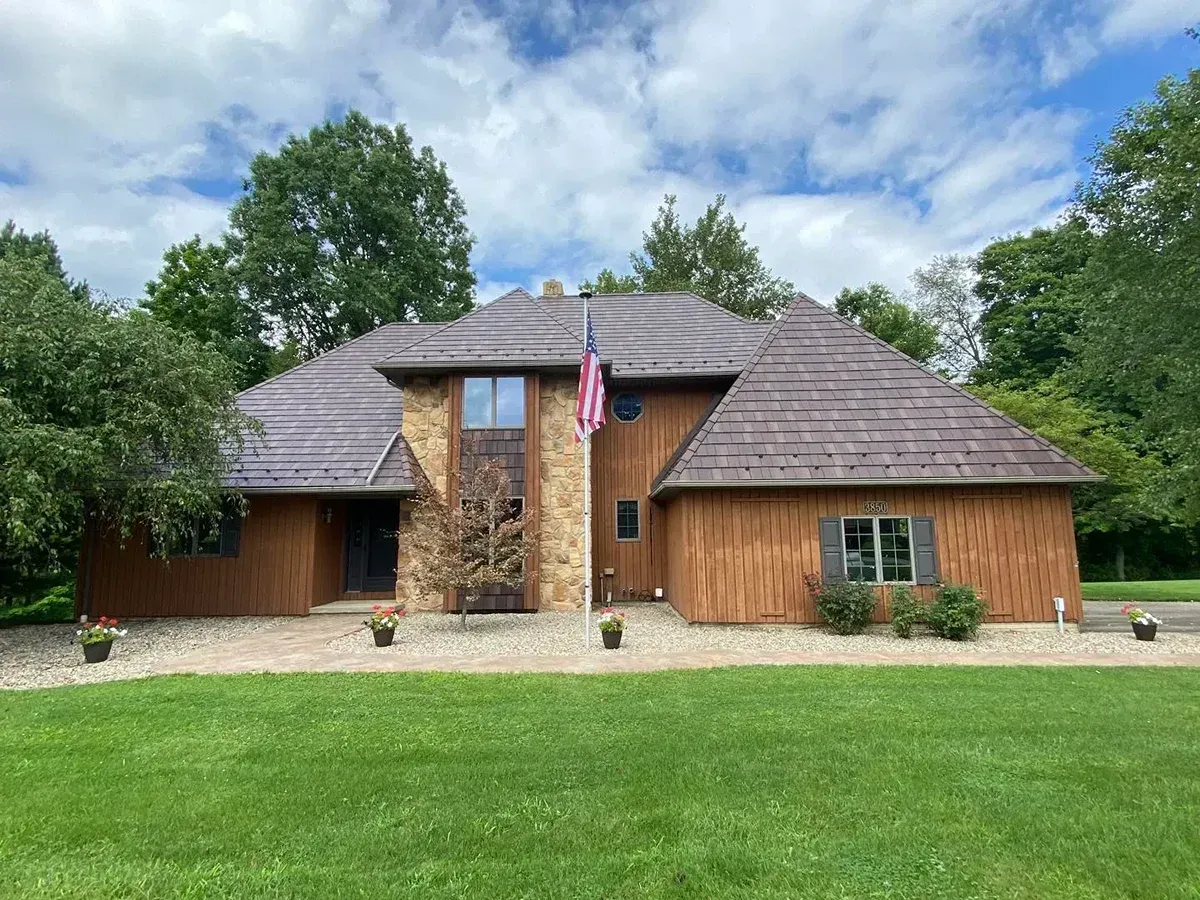 Brown house with stone accents, wood siding, and flag on a green lawn under a cloudy sky.