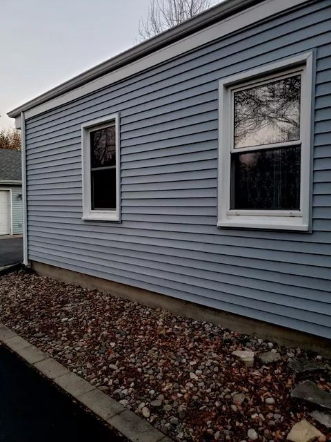 Blue siding with two white-framed windows on a house. Gravel and a dark driveway are below.
