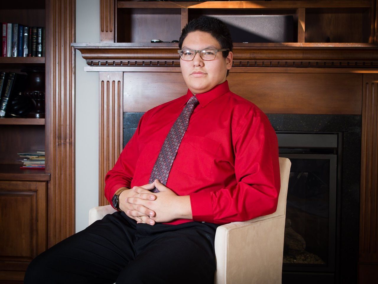 A man in a red shirt and tie is sitting in front of a fireplace.