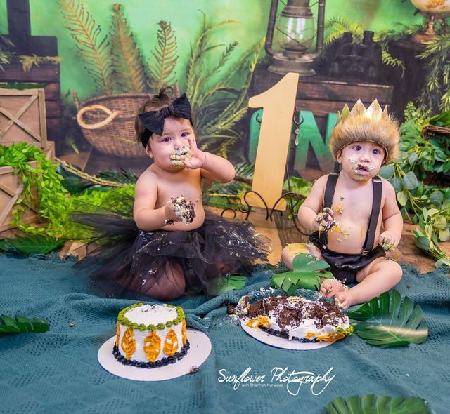 Two babies are sitting on a blanket eating cake.