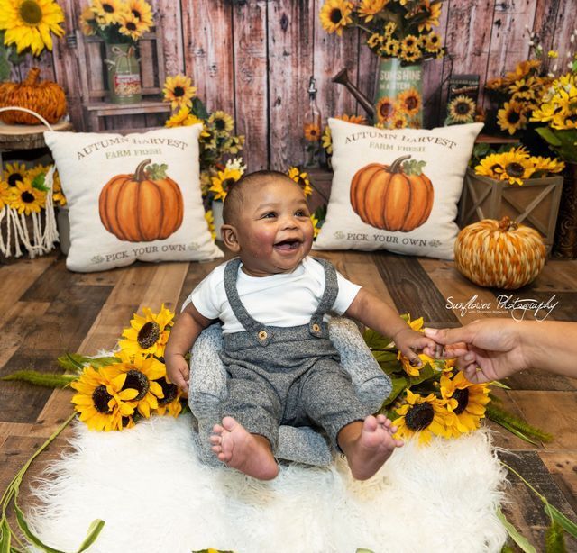 A baby is sitting next to a pillow that says autumn harvest