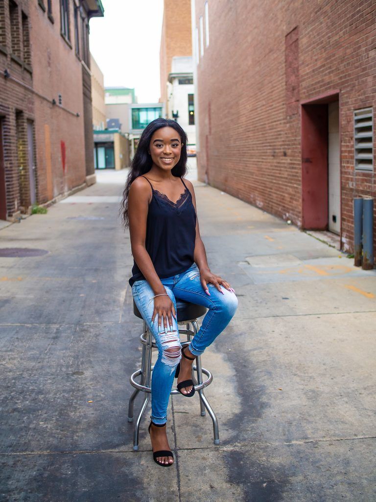 A woman is sitting on a bar stool in an alleyway.