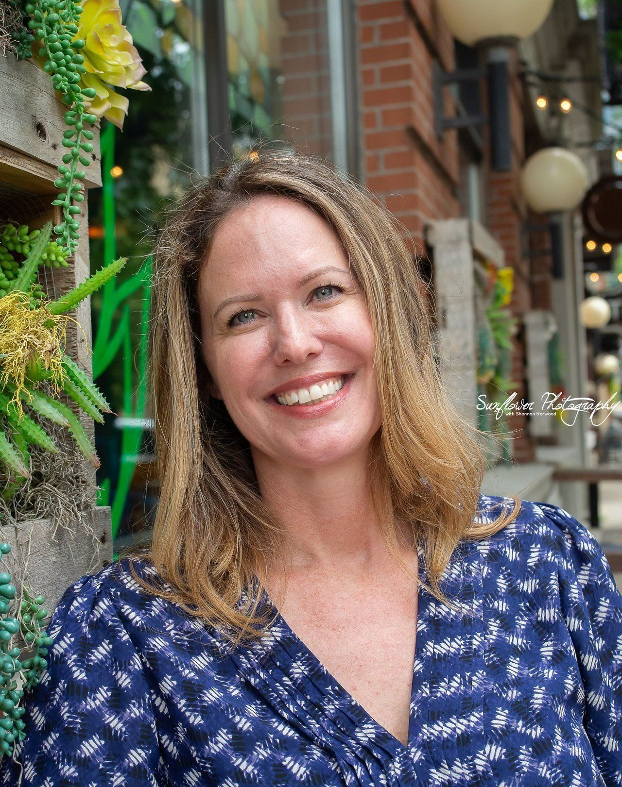 A woman in a blue shirt is smiling in front of a brick building.