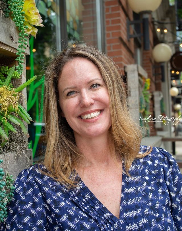A woman in a blue shirt is smiling in front of a brick building.