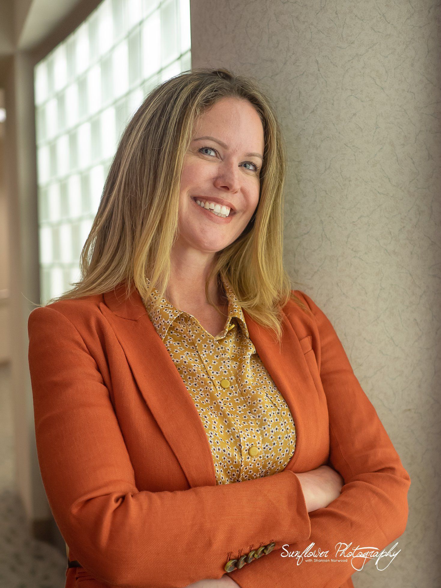 A woman in an orange jacket and yellow shirt is smiling with her arms crossed.