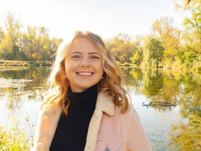 A woman in a pink jacket is smiling in front of a lake.