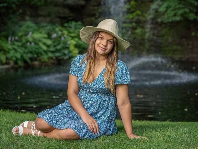 A girl in a blue dress and hat is sitting on the grass in front of a waterfall.